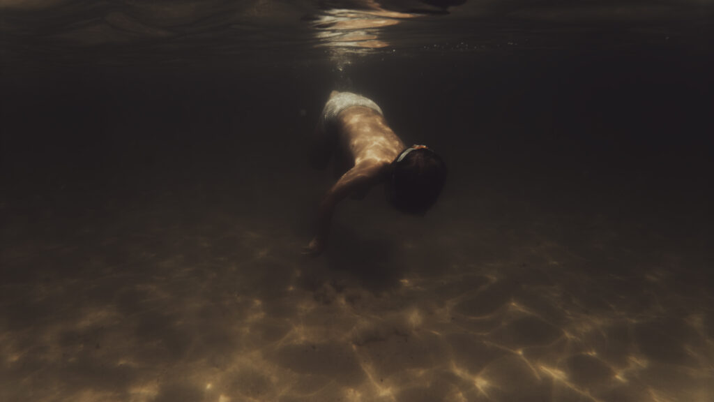 Une personne nage sous l'eau vers le fond sablonneux éclairé par la lumière filtrant depuis la surface.