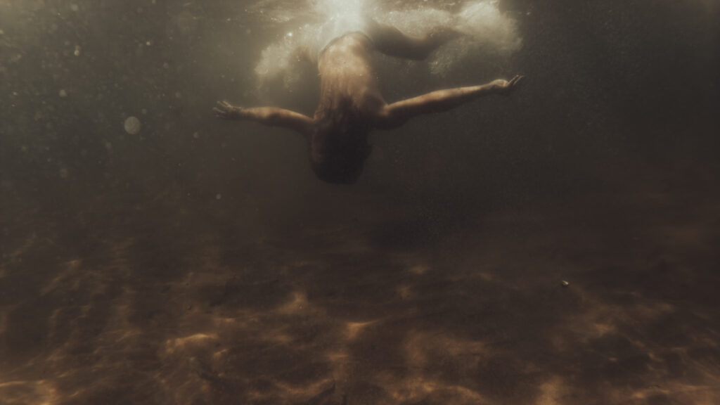 Une personne flotte sous l'eau, bras écartés, baignée par une lumière dorée venant de la surface.