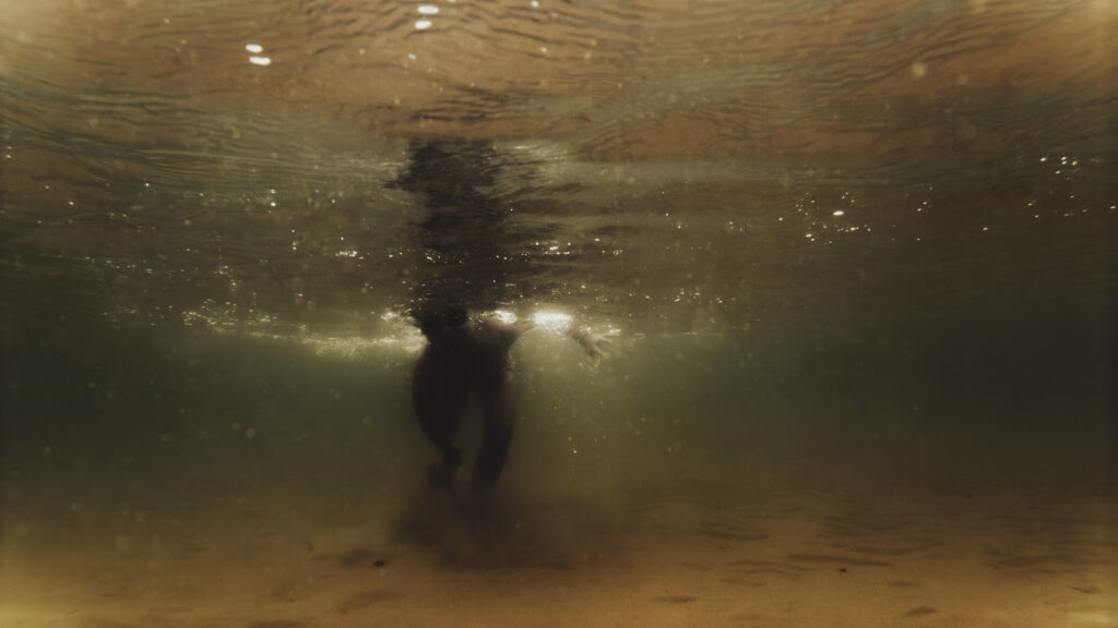 Silhouette d'une personne debout sous l'eau, vue depuis le fond, avec une lumière dorée filtrant à la surface.