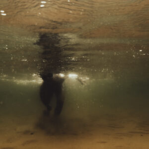 Silhouette d'une personne debout sous l'eau, vue depuis le fond, avec une lumière dorée filtrant à la surface.