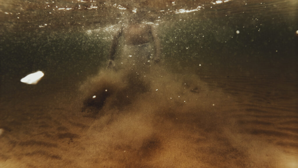 Vue sous-marine d'une personne partiellement visible dans une eau trouble et ambrée, avec du sable en suspension.