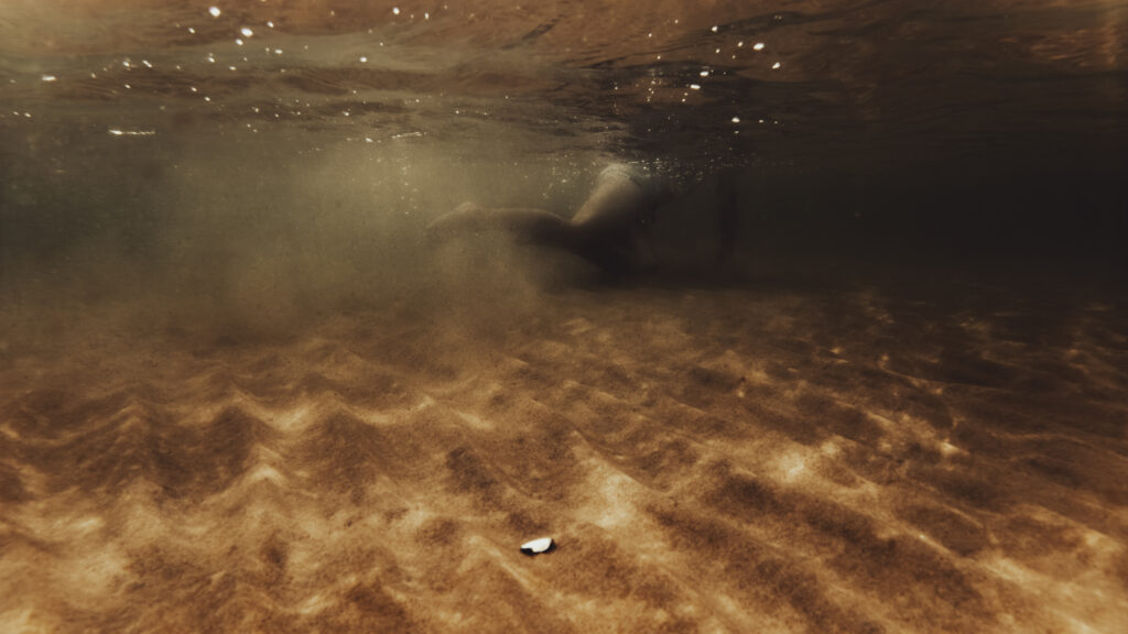 Vue sous-marine des jambes d'une personne nageant au-dessus d'un fond de sable doré, avec la lumière du soleil filtrant à travers la surface de l'eau.