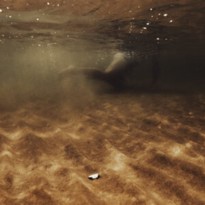 Vue sous-marine des jambes d'une personne nageant au-dessus d'un fond de sable doré, avec la lumière du soleil filtrant à travers la surface de l'eau.