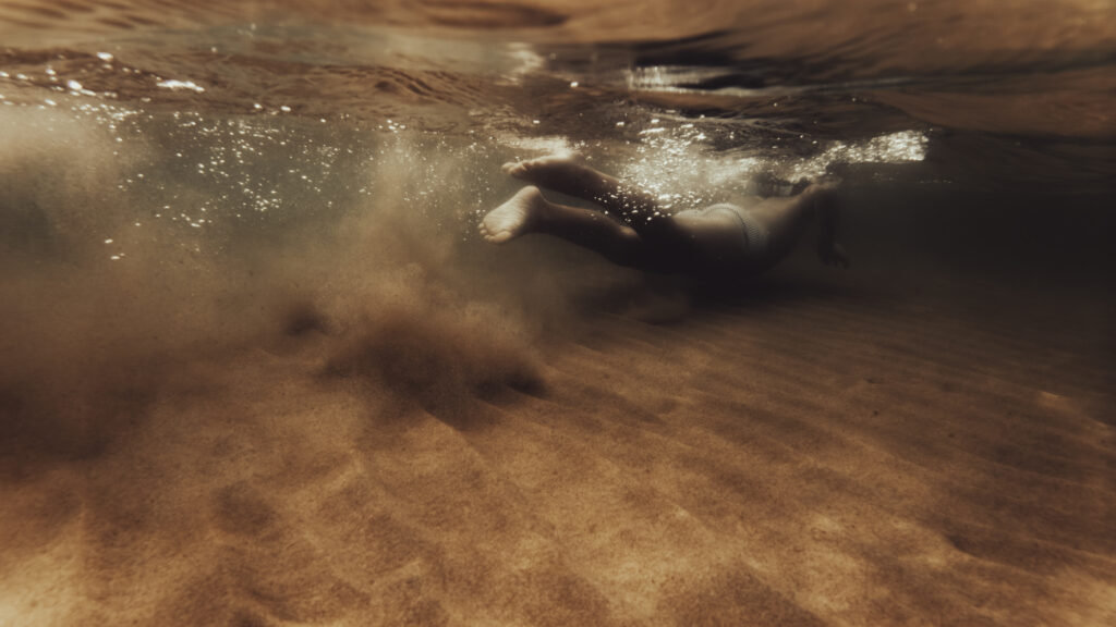 Une personne nage sous l'eau au-dessus d'un fond sableux doré, entourée de bulles lumineuses.