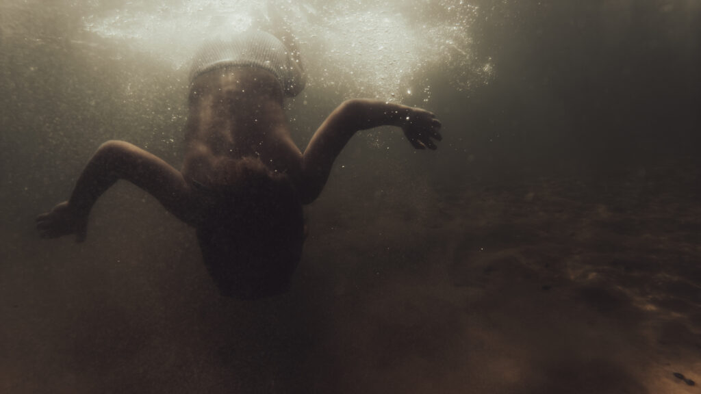 Silhouette d'une personne en bonnet de bain nageant sous l'eau, bras écartés, baignée par une lumière dorée venant de la surface.