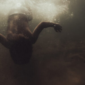 Silhouette d'une personne en bonnet de bain nageant sous l'eau, bras écartés, baignée par une lumière dorée venant de la surface.