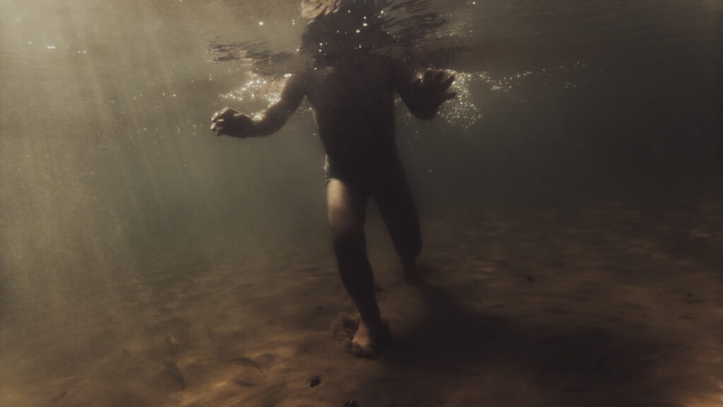 Une personne vue de dos marche sous l'eau, baignée par des rayons de lumière filtrant à travers la surface.