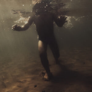 Une personne vue de dos marche sous l'eau, baignée par des rayons de lumière filtrant à travers la surface.