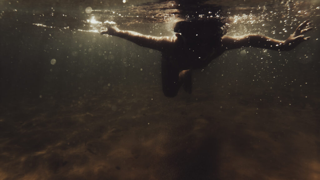 Silhouette d'une personne nageant sous l'eau, bras tendus, traversée par des rayons de lumière dorée.