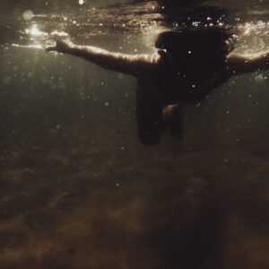 Silhouette d'une personne nageant sous l'eau, bras tendus, traversée par des rayons de lumière dorée.