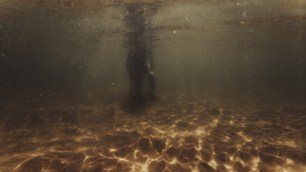 Vue sous-marine d'une silhouette debout dans une eau peu profonde avec un fond de sable éclairé par des reflets dorés.