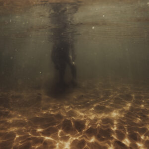 Vue sous-marine d'une silhouette debout dans une eau peu profonde avec un fond de sable éclairé par des reflets dorés.