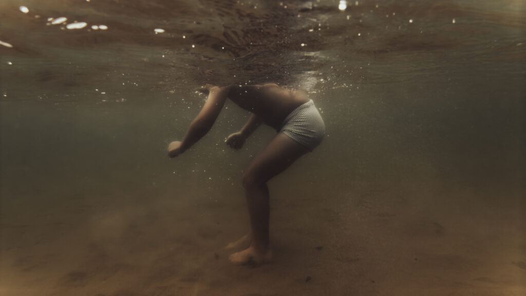 Un enfant en maillot de bain rayé est photographié sous l'eau, penché en avant, la tête hors du cadre à la surface, dans une lumière dorée.