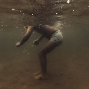 Un enfant en maillot de bain rayé est photographié sous l'eau, penché en avant, la tête hors du cadre à la surface, dans une lumière dorée.