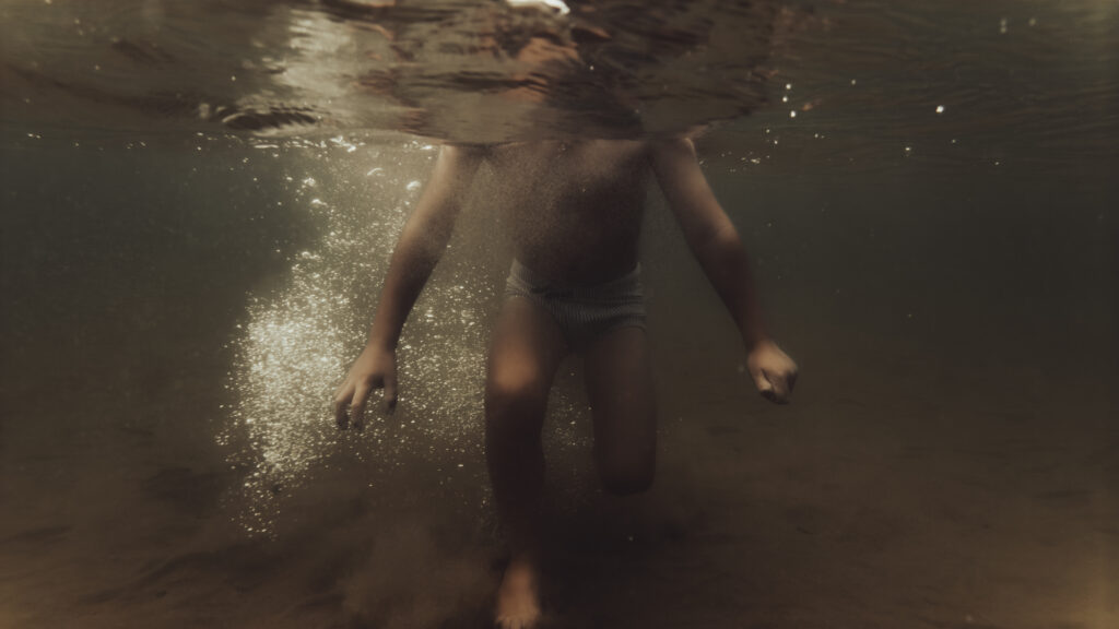 Un enfant en maillot de bain rayé marche sous la surface de l'eau, baigné d'une lumière dorée et entouré de bulles.