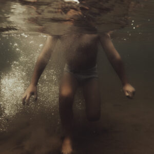 Un enfant en maillot de bain rayé marche sous la surface de l'eau, baigné d'une lumière dorée et entouré de bulles.