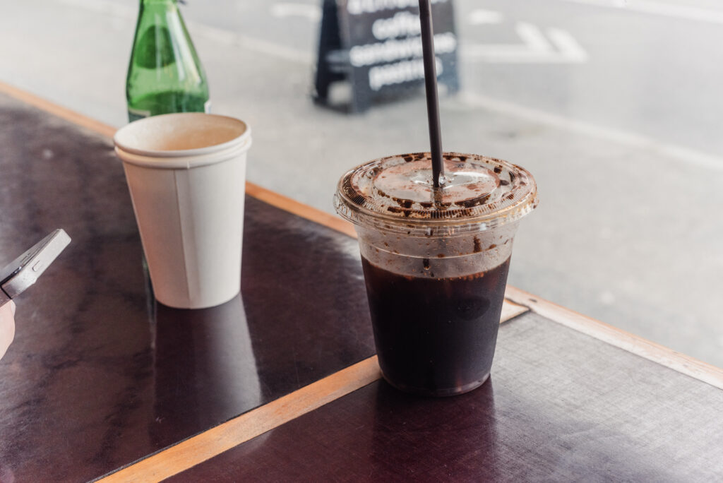 Un café glacé dans un gobelet en plastique avec une paille noire, posé sur une table en bois à côté d'un gobelet en carton et d'une bouteille d'eau gazeuse verte, en terrasse.