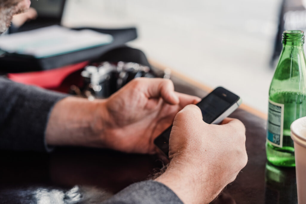 Gros plan sur des mains tenant un smartphone à une table, avec une bouteille en verre vert et une boisson glacée en arrière-plan.