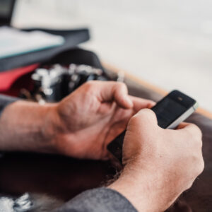 Gros plan sur des mains tenant un smartphone à une table, avec une bouteille en verre vert et une boisson glacée en arrière-plan.