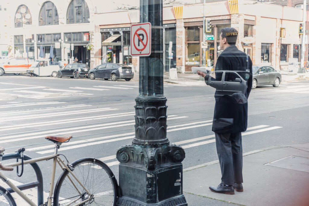 Un homme en costume attend au bord d'un trottoir dans une rue commerçante, un document à la main, avec un vélo attaché à proximité.