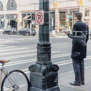 Un homme en costume attend au bord d'un trottoir dans une rue commerçante, un document à la main, avec un vélo attaché à proximité.