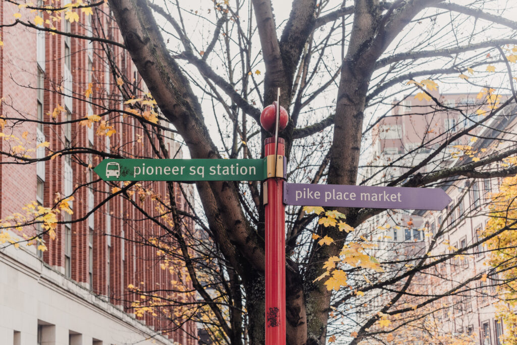 Un panneau de signalisation rouge indiquant Pioneer Sq Station et Pike Place Market, entouré des branches d'un arbre aux feuilles d'automne devant des immeubles en briques.