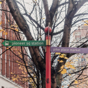 Un panneau de signalisation rouge indiquant Pioneer Sq Station et Pike Place Market, entouré des branches d'un arbre aux feuilles d'automne devant des immeubles en briques.