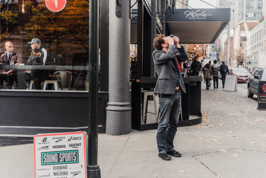 Un homme en veste grise prend une photo en visant vers le haut, debout sur un trottoir animé bordé de commerces.
