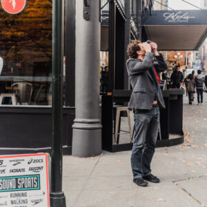 Un homme en veste grise prend une photo en visant vers le haut, debout sur un trottoir animé bordé de commerces.