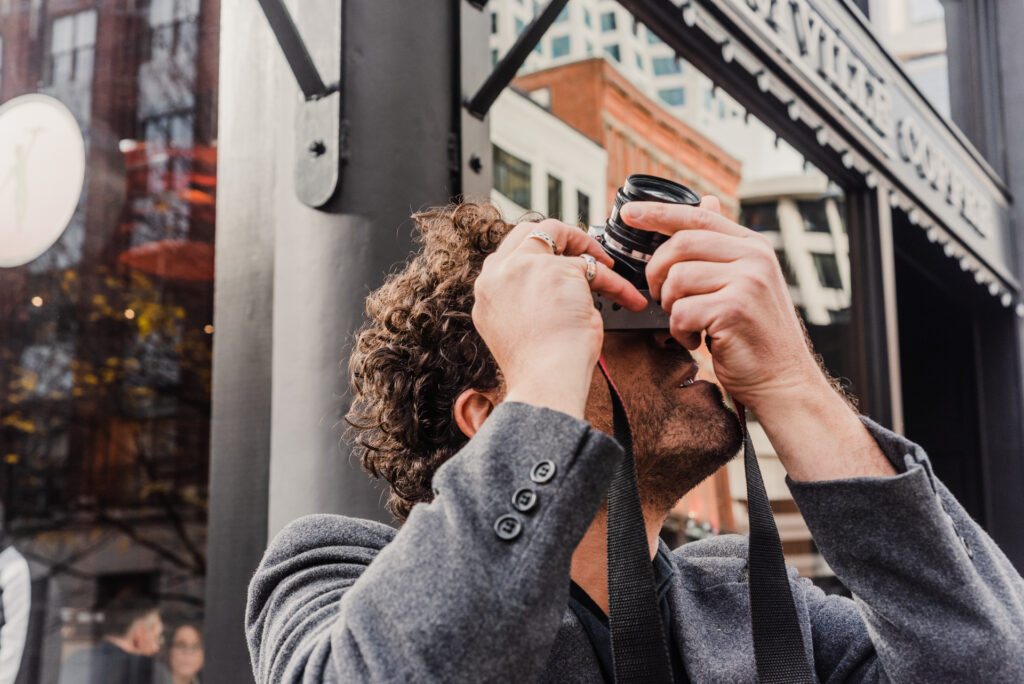 Un homme aux cheveux bouclés lève un appareil photo pour photographier un bâtiment dans une rue animée.