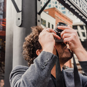 Un homme aux cheveux bouclés lève un appareil photo pour photographier un bâtiment dans une rue animée.