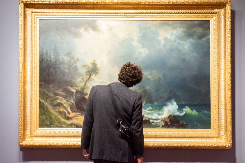 Une personne vue de dos, appareil photo à l'épaule, contemple une grande peinture de paysage marin dans un cadre doré.