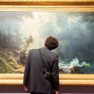 Une personne vue de dos, appareil photo à l'épaule, contemple une grande peinture de paysage marin dans un cadre doré.