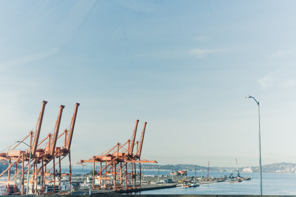 Des grues à conteneurs orange se dressent le long d'un quai portuaire, avec une baie et des collines en arrière-plan sous un ciel dégagé.
