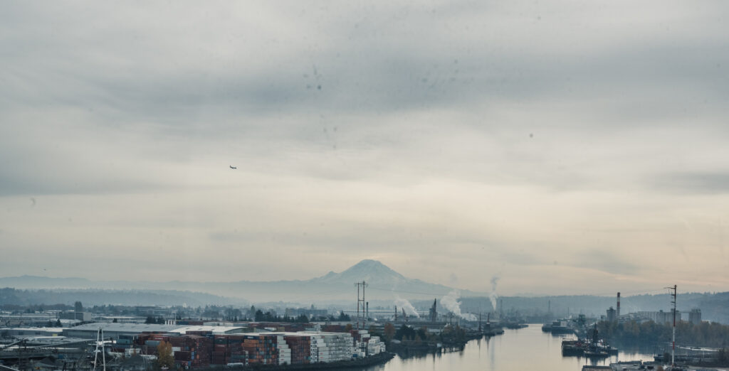 Vue d'un port industriel avec des conteneurs empilés le long d'une rivière, des cheminées fumantes et un volcan enneigé visible sous un ciel brumeux.