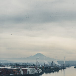 Vue d'un port industriel avec des conteneurs empilés le long d'une rivière, des cheminées fumantes et un volcan enneigé visible sous un ciel brumeux.