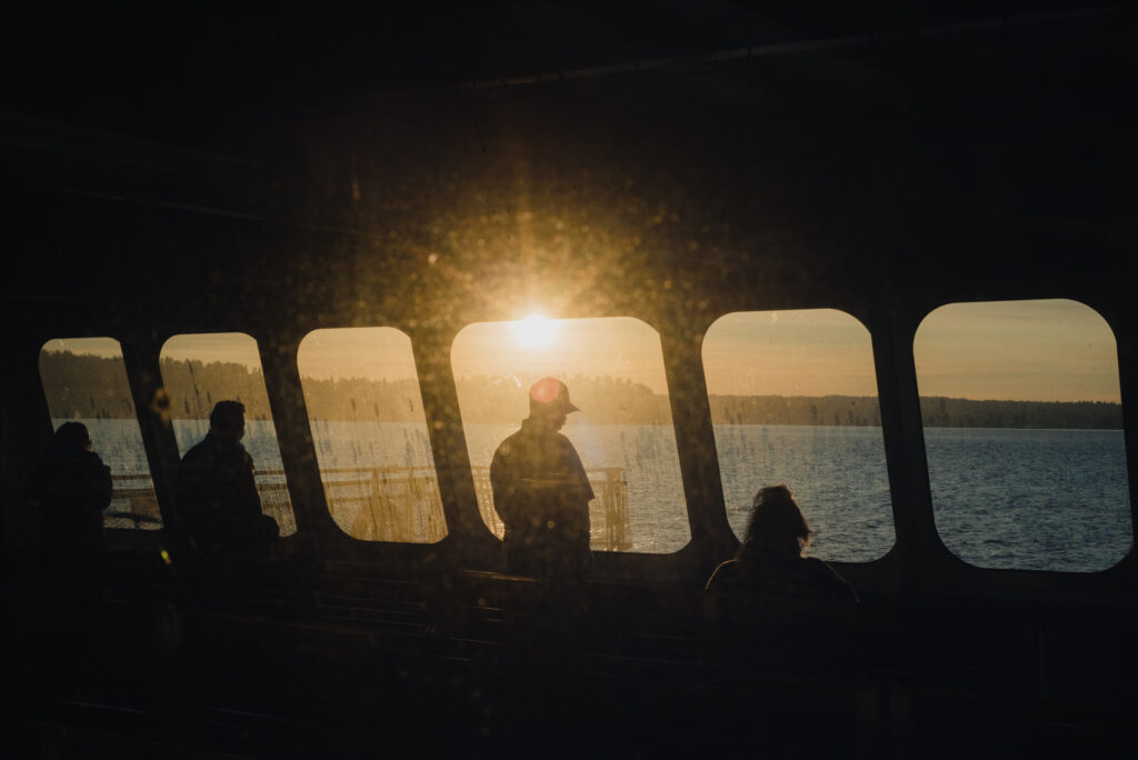 Des passagers en silhouette regardent un coucher de soleil doré depuis les fenêtres d'un ferry face à un lac.