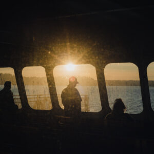 Des passagers en silhouette regardent un coucher de soleil doré depuis les fenêtres d'un ferry face à un lac.