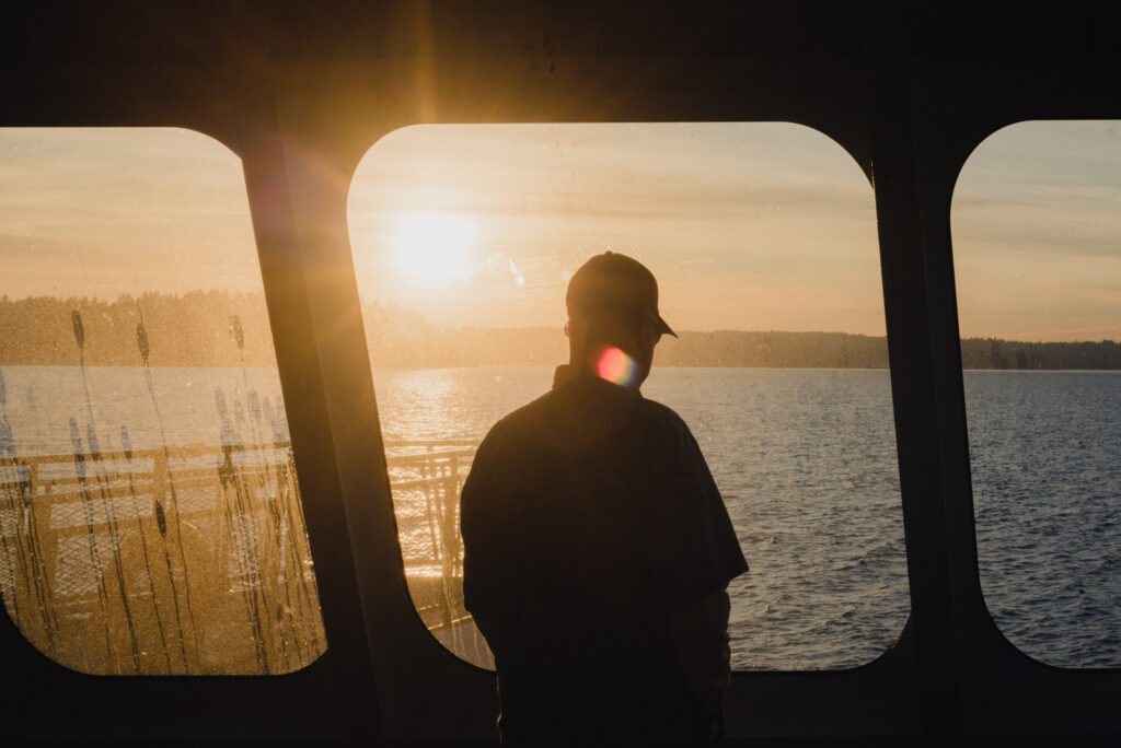 Silhouette d'une personne portant une casquette, regardant un lac par la fenêtre d'un bateau au coucher du soleil