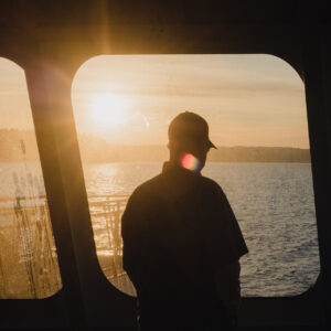 Silhouette d'une personne portant une casquette, regardant un lac par la fenêtre d'un bateau au coucher du soleil