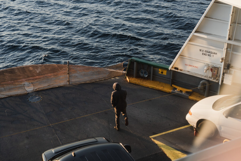 Une personne en veste à capuche marche seule sur le pont voiture d'un ferry, face à la mer.
