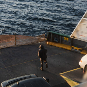 Une personne en veste à capuche marche seule sur le pont voiture d'un ferry, face à la mer.