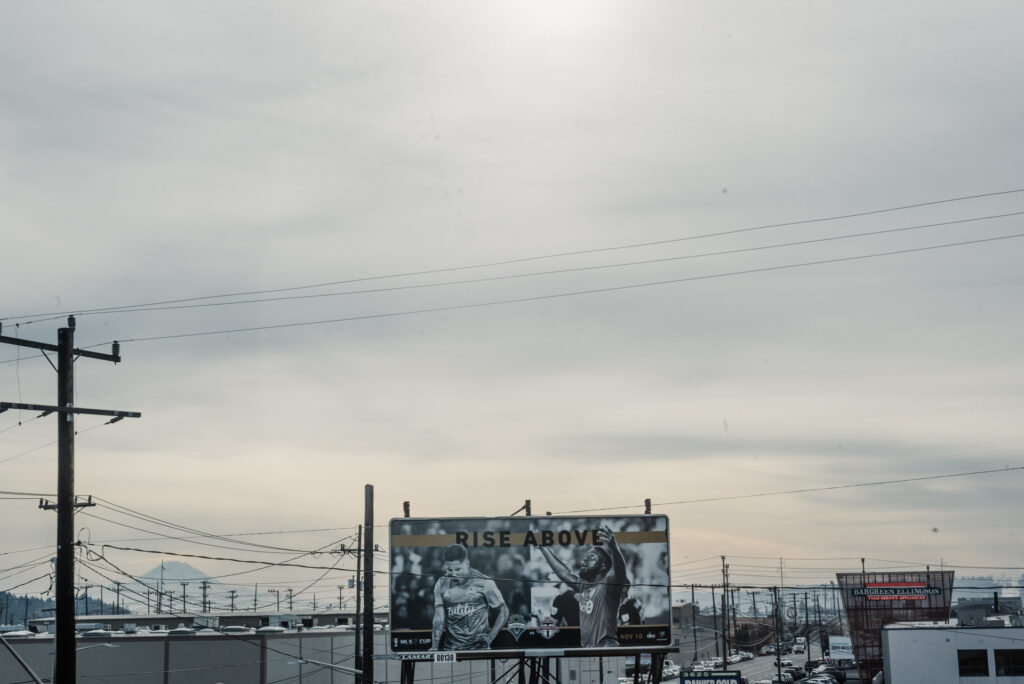 Un grand panneau d'affichage publicitaire pour un match de football se dresse dans une zone industrielle sous un ciel gris nuageux, avec une montagne visible au loin.
