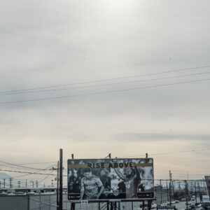 Un grand panneau d'affichage publicitaire pour un match de football se dresse dans une zone industrielle sous un ciel gris nuageux, avec une montagne visible au loin.
