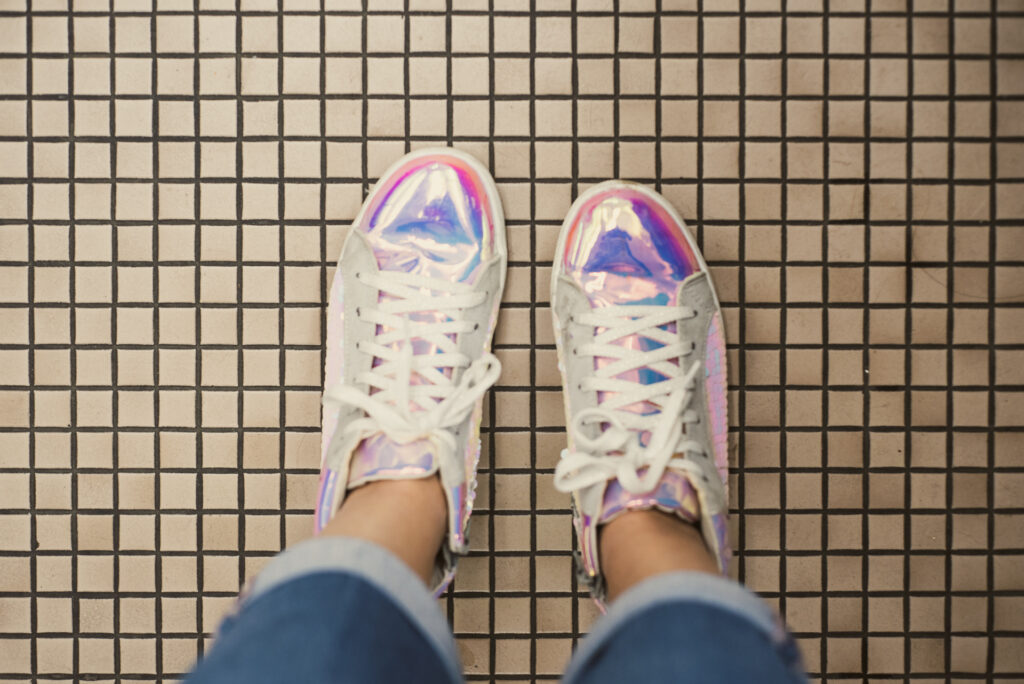 Vue du dessus de deux baskets holographiques portées avec un jean sur un sol carrelé quadrillé.