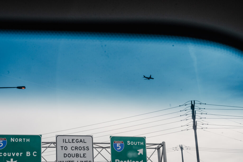 Un avion vole dans un ciel bleu au-dessus de panneaux d'autoroute indiquant les directions vers Vancouver BC et Portland, vue depuis l'intérieur d'un véhicule.