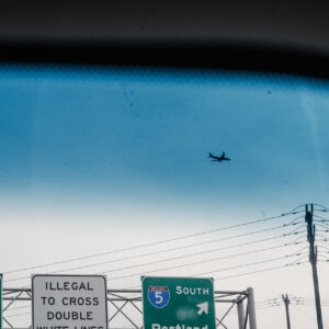 Un avion vole dans un ciel bleu au-dessus de panneaux d'autoroute indiquant les directions vers Vancouver BC et Portland, vue depuis l'intérieur d'un véhicule.