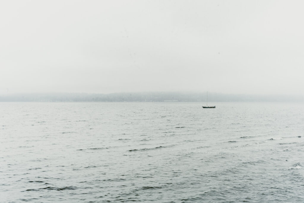 Un voilier seul flotte sur une mer grise, face à une côte boisée voilée par la brume.