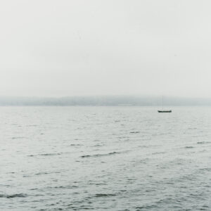 Un voilier seul flotte sur une mer grise, face à une côte boisée voilée par la brume.