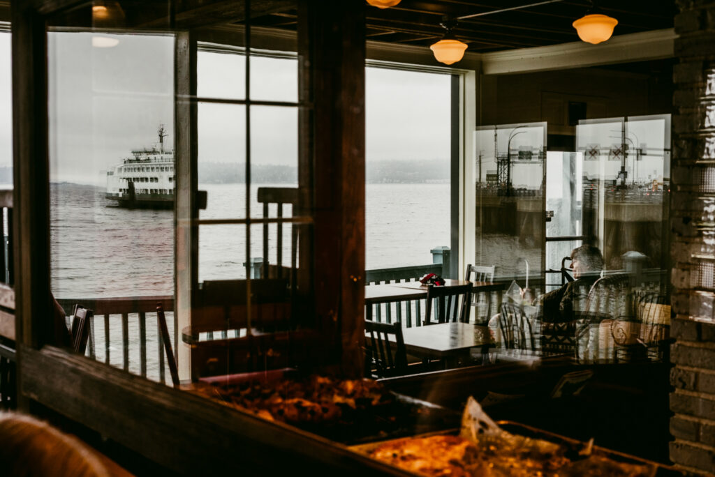 Une personne assise près d'une fenêtre de restaurant regarde un ferry naviguer sur une mer brumeuse.